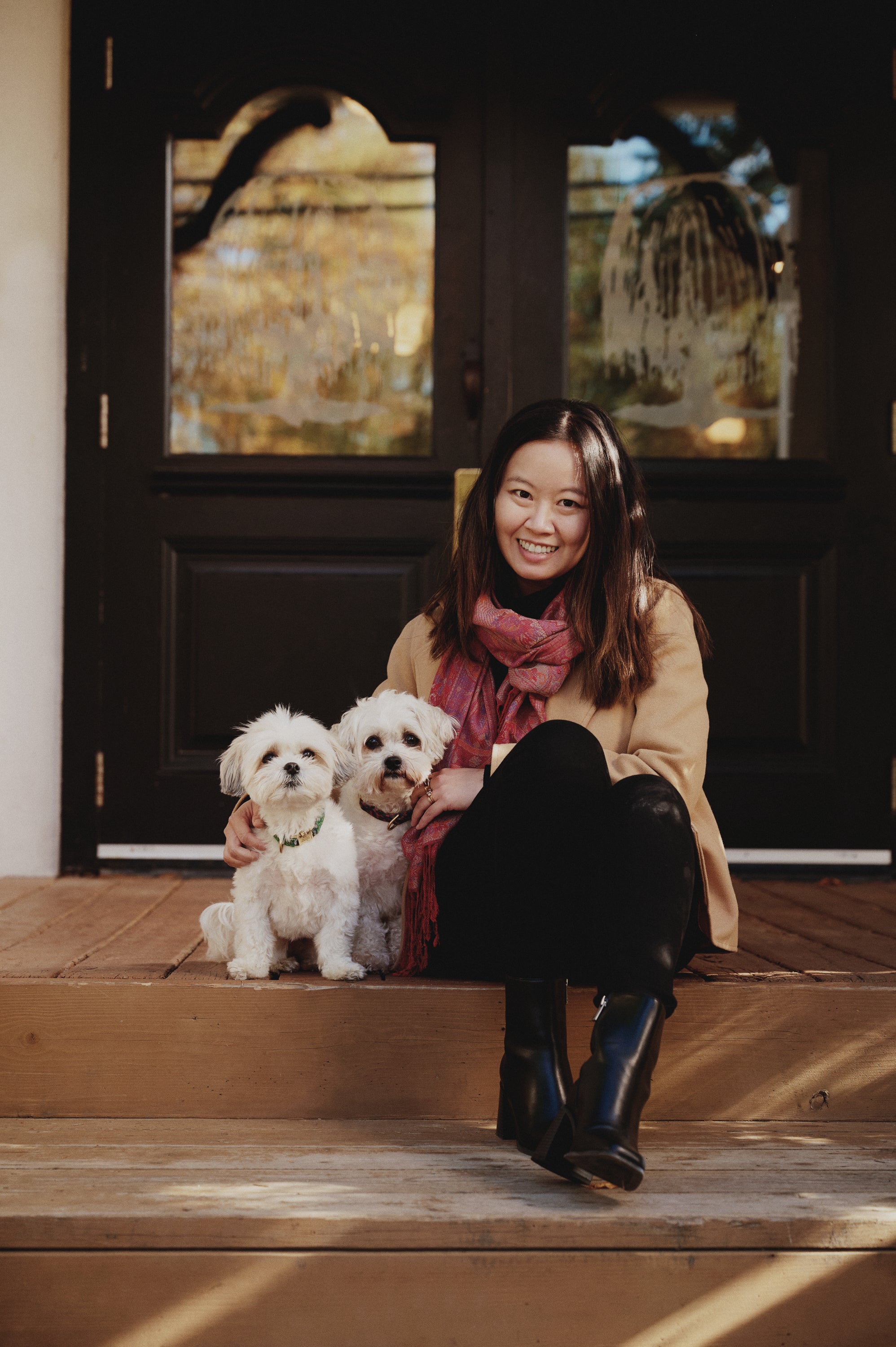 Purely Golden Foods founder Irene Ou sitting on front porch steps with her two white Maltese dogs, smiling outdoors in autumn light